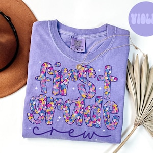 May include: A violet-colored t-shirt with the words "First Grade Crew" in a floral design. The shirt has a crew neck and short sleeves. A gold necklace is draped across the shirt. A brown hat is in the background.