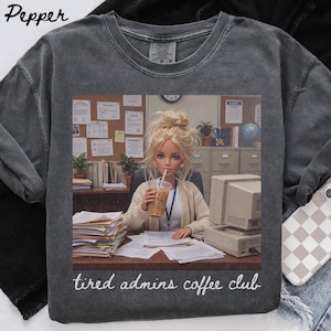 Op de afbeelding: Een antracietgrijs t-shirt met ronde hals en een grafische print van een blonde pop in een beige vest, zittend aan een bureau met een computer, papieren en een koffiebeker. De tekst "tired admins coffee club" is in het wit onder de print gedrukt.