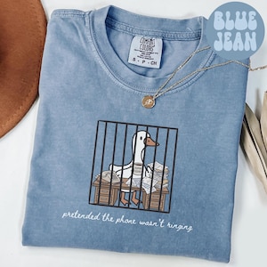 May include: A blue short-sleeve t-shirt featuring a graphic of a white goose sitting at a desk inside a cage, surrounded by papers and a telephone. The text "pretended the phone wasn't ringing" is printed below the graphic in white script.