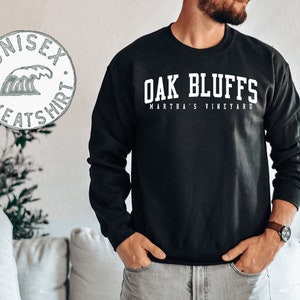 Op de afbeelding: Zwart sweatshirt met witte tekst die "OAK BLUFFS MARTHA'S VINEYARD" luidt. Een witte cirkel met een golfgrafiek en de tekst "UNISEX SWEATSHIRT" is op de achtergrond.