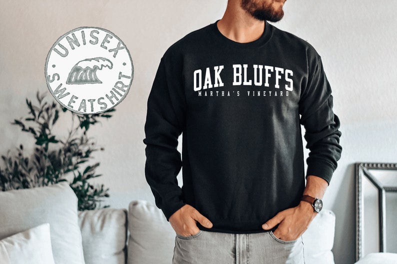 Op de afbeelding: Zwart sweatshirt met witte tekst die "OAK BLUFFS MARTHA'S VINEYARD" luidt. Een witte cirkel met een golfgrafiek en de tekst "UNISEX SWEATSHIRT" is op de achtergrond.