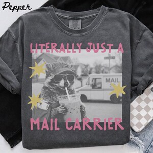 Könnte beinhalten: Ein anthrazitfarbenes T-Shirt mit einer schwarz-weißen Grafik eines Waschbären, der eine Baseballkappe und eine Sonnenbrille trägt und ein Getränk mit Strohhalm hält. Der Text LITERALLY JUST A MAIL CARRIER ist in Pink aufgedruckt.