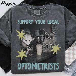 May include: A charcoal gray t-shirt featuring a graphic of an opossum, a skunk, and a raccoon in an eye exam setting. The text reads "SUPPORT YOUR LOCAL OPTOMETRISTS" in light blue. The animals are depicted with optometry tools and sunglasses.