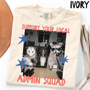 May include: An ivory t-shirt featuring a graphic of an opossum, a skunk, and a raccoon in an office setting. The text reads "SUPPORT YOUR LOCAL ADMIN SQUAD". The raccoon wears sunglasses and holds a telephone receiver. Blue star shapes accent the graphic.