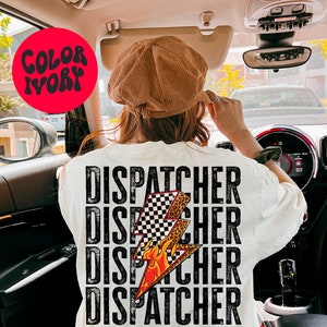 以下が含まれることがあります： 白いTシャツに、黒い文字で「DISPATCHER」という単語が4回繰り返されています。シャツの中央には、炎がついた黒と白の市松模様の稲妻が描かれています。