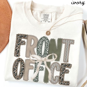 May include: An ivory t-shirt featuring the text "FRONT OFFICE" in a decorative style. The letters incorporate patterns like leopard print and stripes, with a white bow detail. A gold necklace with a pendant is draped across the shirt.