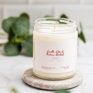 a white candle in a jar with a label that reads "Smells like a Badass Dentist", placed on a small stone coaster.