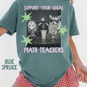 May include: A blue spruce colored t-shirt featuring a graphic of a possum, a skunk, and a raccoon wearing sunglasses at a desk with a calculator, a drink, and a ruler. The text reads SUPPORT YOUR LOCAL MATH TEACHERS with light green star accents.