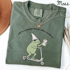 Op de afbeelding: Een mosgroen t-shirt met een afbeelding van een groene kikker met een puntmuts op een step, die een klembord vasthoudt. De tekst "off to commit front office foolery" staat boven de kikker. Het shirt heeft een ronde halslijn.