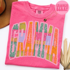 May include: A pink t-shirt with the word "GRANDMA" in a patchwork design. The letters are filled with various paisley patterns in shades of green, orange, pink, and purple. The shirt has a crew neck and a small tag that says "COMFORT COLORS". A gold necklace is also visible.