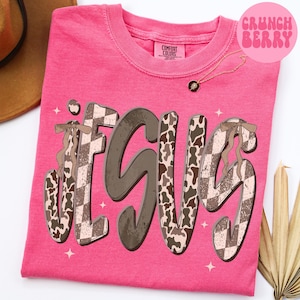 May include: A pink Comfort Colors t-shirt with the word "JESUS" in a unique font. The letters have a mix of brown, cow print, and checkered patterns. The shirt also features a small gold necklace and the text "Crunch Berry" in a pink circle.