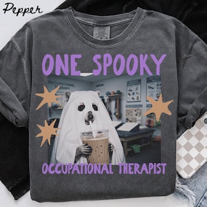 May include: A charcoal gray t-shirt featuring a graphic of a bear wearing a white ghost costume and holding an iced coffee. The text reads ONE SPOOKY OCCUPATIONAL THERAPIST in purple letters, accented by tan star shapes on a background of a therapy room.