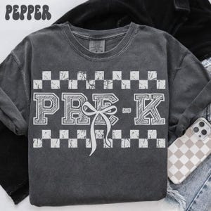 May include: A dark grey t-shirt with a white checkered pattern and the text "PRE-K" in a white block font with a white bow.