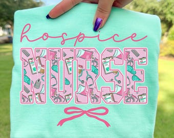 May include: A light turquoise t-shirt with the words "hospice NURSE" in pink script and block letters. The word "NURSE" is filled with medical-themed illustrations. A pink bow is below the text. The brand name "Island Reef" is at the bottom.