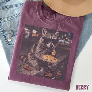 May include: A berry-colored crew neck t-shirt featuring a graphic print of a raccoon wearing black sunglasses and eating a deviled egg from a small plate. The shirt has a Comfort Colors label at the inner neckline with the text XL-TG-EG.