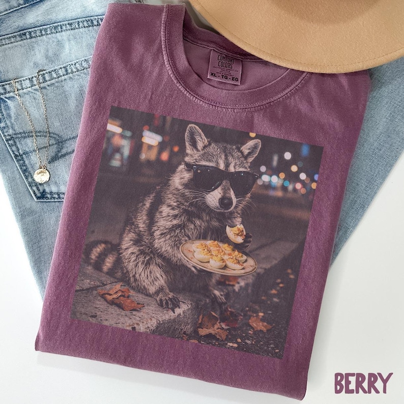 May include: A berry-colored crew neck t-shirt featuring a graphic print of a raccoon wearing black sunglasses and eating a deviled egg from a small plate. The shirt has a Comfort Colors label at the inner neckline with the text XL-TG-EG.