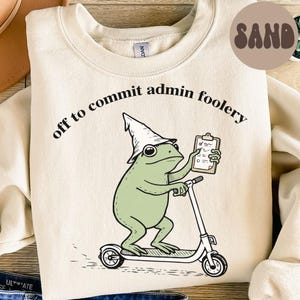 May include: A sand-colored crewneck sweatshirt featuring a graphic of a green frog wearing a pointed hat, riding a scooter, and holding a clipboard. The text above the frog reads, off to commit admin foolery, in a black, curved font.