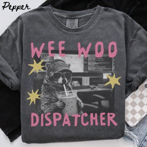 Puede incluir: Una camiseta gris carbón de cuello redondo con un gráfico de un mapache con auriculares y gafas de sol sosteniendo una bebida. El texto WEE WOO DISPATCHER aparece en letras rosas junto a estrellas amarillas sobre un fondo de oficina en blanco y negro.