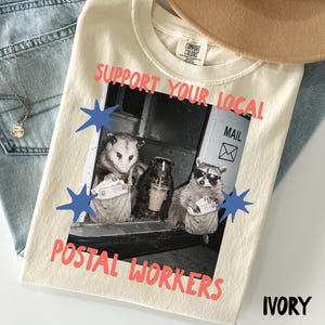 May include: An ivory t-shirt featuring a graphic of an opossum, a skunk, and a raccoon dressed as postal workers. The text reads SUPPORT YOUR LOCAL POSTAL WORKERS. The animals are shown with mail bags and a coffee cup, with blue star accents.