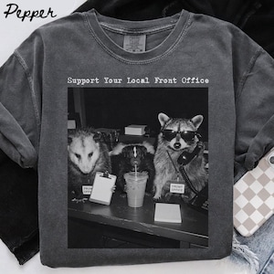 Könnte beinhalten: Ein anthrazitfarbenes T-Shirt mit Rundhalsausschnitt und einer schwarz-weißen Grafik eines Opossums, eines Stinktiers und eines Waschbären an einem Schreibtisch. Der Text darüber lautet Support Your Local Front Office, mit Namensschildern.