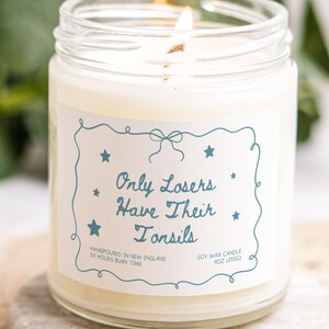 Könnte beinhalten: Eine brennende Sojawachskerze in einem durchsichtigen Glas. Die Kerze hat ein Etikett mit dem Text "Only Losers Have Their Tonsils" in blauer Schrift. Das Etikett enthält auch den Text "Handpoured in New England" und "50 Hours Burn Time".