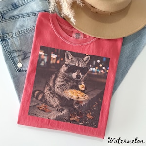 May include: A coral-colored t-shirt featuring a graphic print of a raccoon wearing black sunglasses and holding a plate of deviled eggs. The shirt has a crew neckline and a Comfort Colors brand label on the inside collar area.