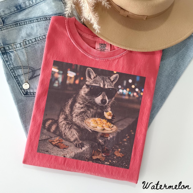 May include: A coral-colored t-shirt featuring a graphic print of a raccoon wearing black sunglasses and holding a plate of deviled eggs. The shirt has a crew neckline and a Comfort Colors brand label on the inside collar area.