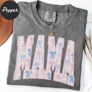 A gray t-shirt with the word "MAMA" printed in pink and blue letters, along with a necklace and a brown hat.