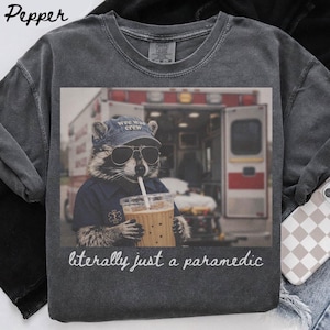 Könnte beinhalten: Ein anthrazitfarbenes T-Shirt mit Rundhalsausschnitt und einer Grafik eines Waschbären, der eine blaue Mütze mit der Aufschrift 'wee woo crew' und ein blaues Hemd mit Stern-Emblem trägt. Er hält einen Becher. Der Text lautet 'literally just a paramedic'.