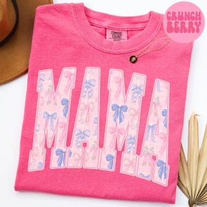 A pink t-shirt with the word "MAMA" printed in large letters, featuring a pattern of pink and blue bows.