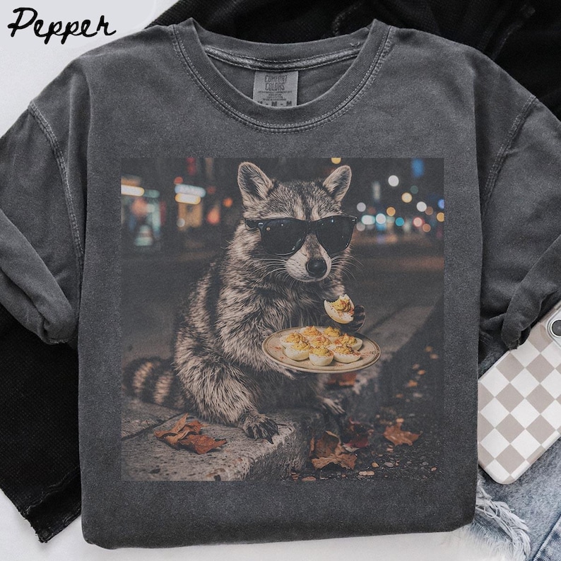 May include: A charcoal gray crew neck t-shirt featuring a graphic print of a raccoon wearing black sunglasses. The raccoon is sitting on a curb at night, holding a plate of deviled eggs and eating one. The word Pepper is printed in the top left corner.