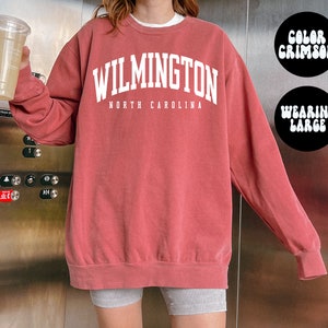 May include: A crimson coloured sweatshirt with the text "WILMINGTON NORTH CAROLINA" in white. The model is wearing a size large.