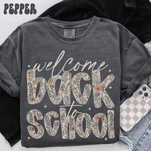 May include: Dark gray long-sleeve shirt with the words "welcome back to school" in a floral design. The word "welcome" is in a cursive font. The letters in "back to school" are filled with a floral pattern of pink, orange, and green.