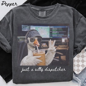 Puede incluir: Una camiseta de cuello redondo gris carbón con el gráfico de un ganso que lleva una gorra, gafas de sol y auriculares, sosteniendo un vaso de plástico con pajita y haciendo el signo de la paz. El texto debajo dice, just a silly dispatcher.