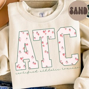 May include: A sand-coloured sweatshirt featuring the letters "ATC" in a collegiate font. The letters are filled with a strawberry pattern and outlined in green. The words "certified athletic trainer" are printed below the letters.