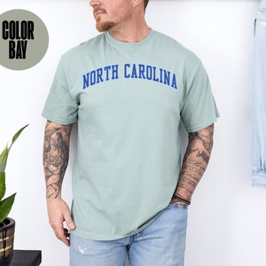 May include: A light green t-shirt with the text "NORTH CAROLINA" in blue arched lettering. The shirt is a solid color and has short sleeves. The shirt is made of a soft, comfortable material.