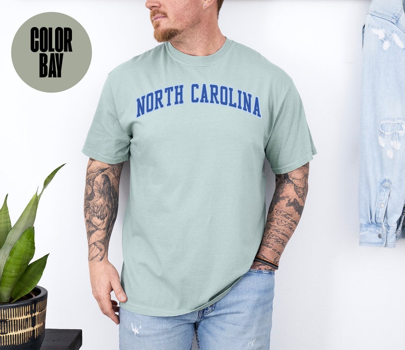 May include: A light green t-shirt with the text "NORTH CAROLINA" in blue arched lettering. The shirt is a solid color and has short sleeves. The shirt is made of a soft, comfortable material.