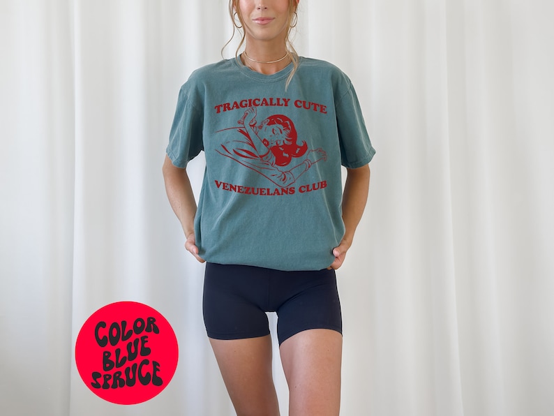 May include: A teal blue t-shirt with a red graphic of a woman lying down with the text "Tragically Cute" and "Venezuelans Club". The shirt is being worn by a person with long brown hair.