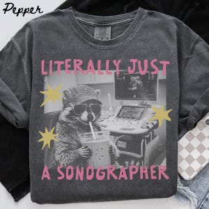 May include: A charcoal gray t-shirt featuring a graphic of a raccoon wearing a baseball cap and sunglasses while holding a drink with a straw. The text reads LITERALLY JUST A SONOGRAPHER in pink letters, accompanied by yellow star shapes.