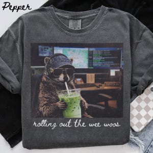 Op de afbeelding: Een antracietgrijs T-shirt met ronde hals en een afbeelding van een wasbeer met een spijkerpet en zonnebril, die een headset draagt en een groen ijsdrankje vasthoudt. De tekst "rolling out the wee woos" staat onder de afbeelding gedrukt.