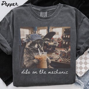 Peut inclure: Un t-shirt à col rond gris anthracite avec une impression graphique représentant un raton laveur portant une casquette en jean et des lunettes de soleil, tenant un café glacé. Le texte "dibs on the mechanic" est écrit en écriture cursive blanche.