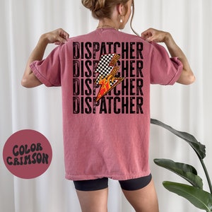 以下が含まれることがあります： 黒文字で「DISPATCHER」という単語が4回繰り返された、クリムゾンカラーのTシャツ。チェッカー柄と炎がついたスタイリッシュな稲妻のグラフィックがTシャツの中央に配置されています。