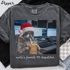 Op de afbeelding: Een antracietgrijs t-shirt met een afbeelding van een wasbeer die een rode kerstmuts en een zonnebril draagt, terwijl deze een drankje met een rietje vasthoudt dat is omwikkeld met gekleurde kerstverlichting. De tekst luidt "santa's favorite 911 dispatcher."