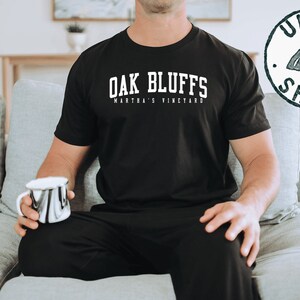 May include: A black t-shirt with white text that reads "Oak Bluffs Martha's Vineyard". The person is wearing black pants and sitting on a gray couch.