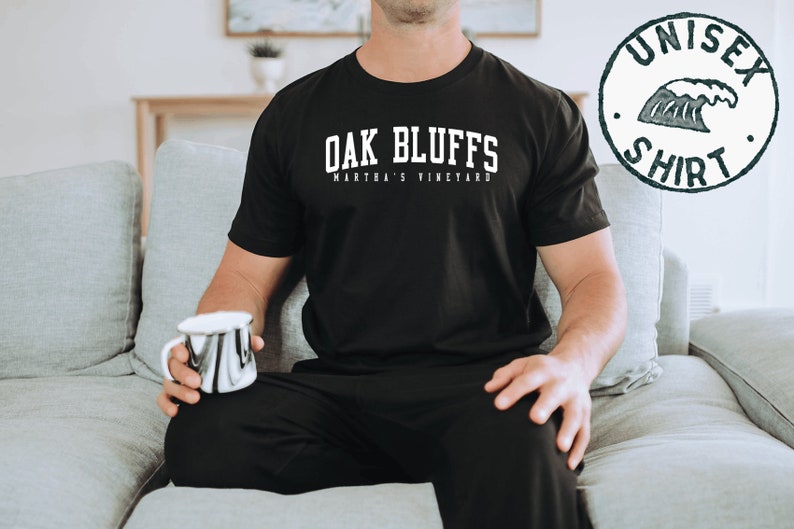 May include: A black t-shirt with white text that reads "Oak Bluffs Martha's Vineyard". The person is wearing black pants and sitting on a gray couch.