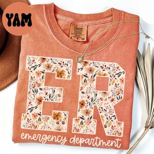 May include: A coral-colored t-shirt with the letters "ER" in a floral design, with the words "emergency department" below. A gold necklace with a pendant is draped on the shirt.