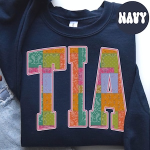 Puede incluir: Sudadera azul marino con la palabra "TIA" en letras grandes y coloridas. Las letras están rellenas con un diseño de patchwork de estampados de cachemira en tonos rosa, naranja, verde y morado. La palabra "NAVY" está en blanco, con fuente redondeada en la esquina superior derecha.