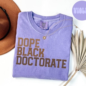 Puede incluir: Una camiseta morada con el texto "Dope Black Doctorate" impreso en marrón en la parte delantera. La camiseta está doblada y colocada sobre una superficie blanca.