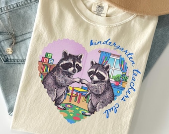 May include: An ivory t-shirt featuring a heart-shaped graphic of two raccoons in a classroom setting. The design includes a bookshelf with ABC blocks, a window, and colorful floor mats. The text reads kindergarten teachers club in blue script.