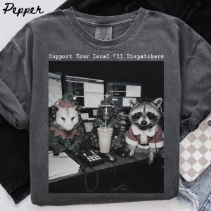 Puede incluir: Una camiseta gris carbón con un estampado gráfico de una zarigüeya, una mofeta y un mapache con ropa festiva y auriculares en un escritorio de despacho. El texto "Support Your Local 911 Dispatchers" está impreso sobre el gráfico.